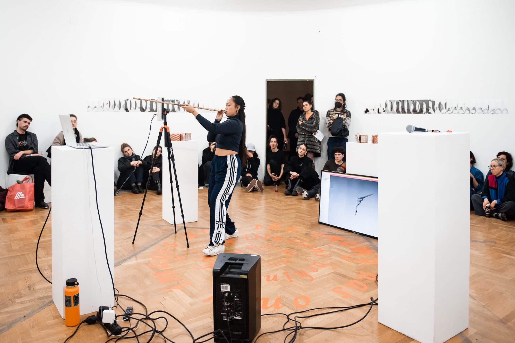 Artist has her mouth to a blowgun and is standing in front of a 3D scanning machine in the middle of an art gallery