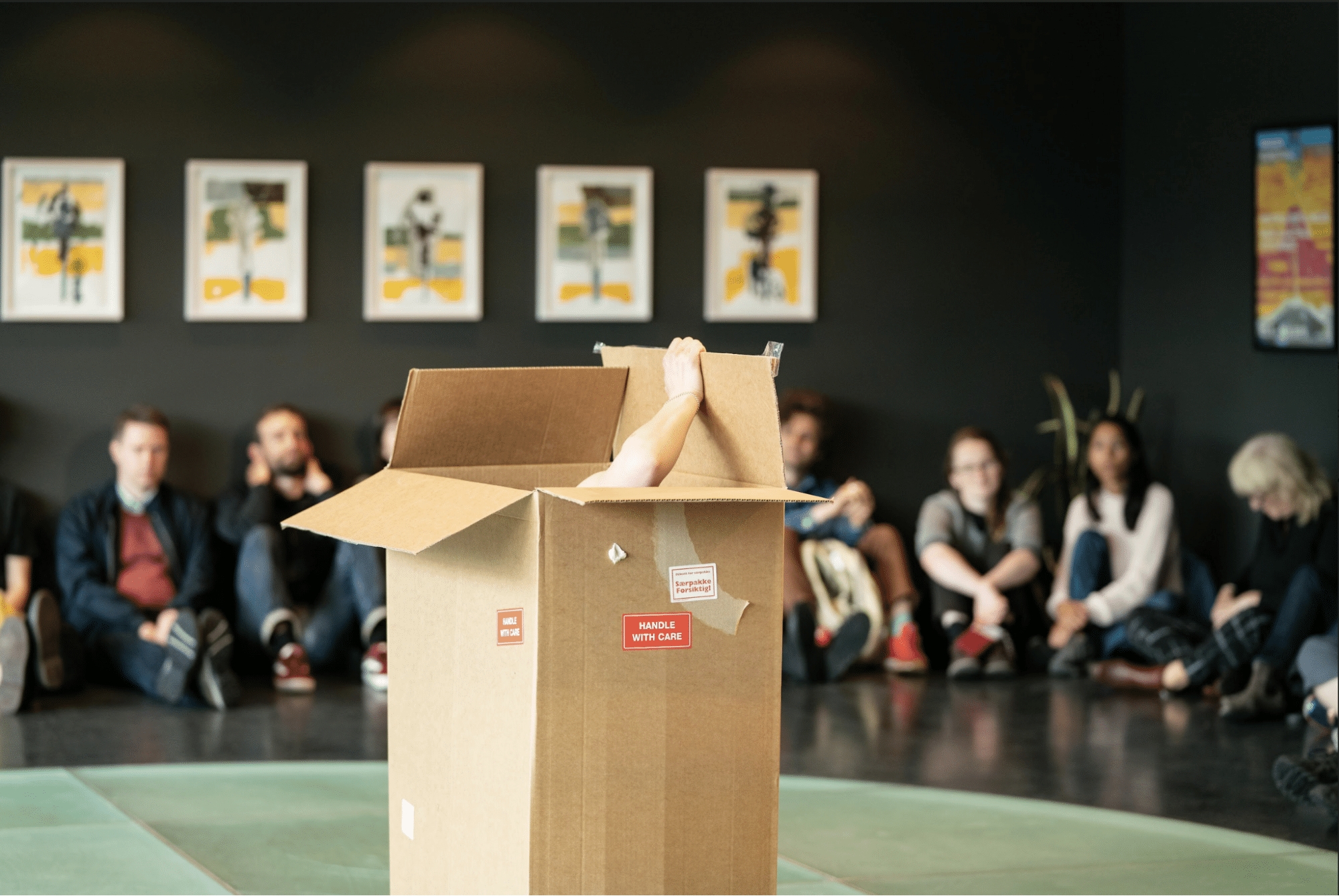 There is a large brown cardboard box in the foreground with the top open and a light skinned human arm extending out of it, opening one of the flaps. The background is out of focus and shows an audience of people sitting on the floor of a gray room with art on the walls.