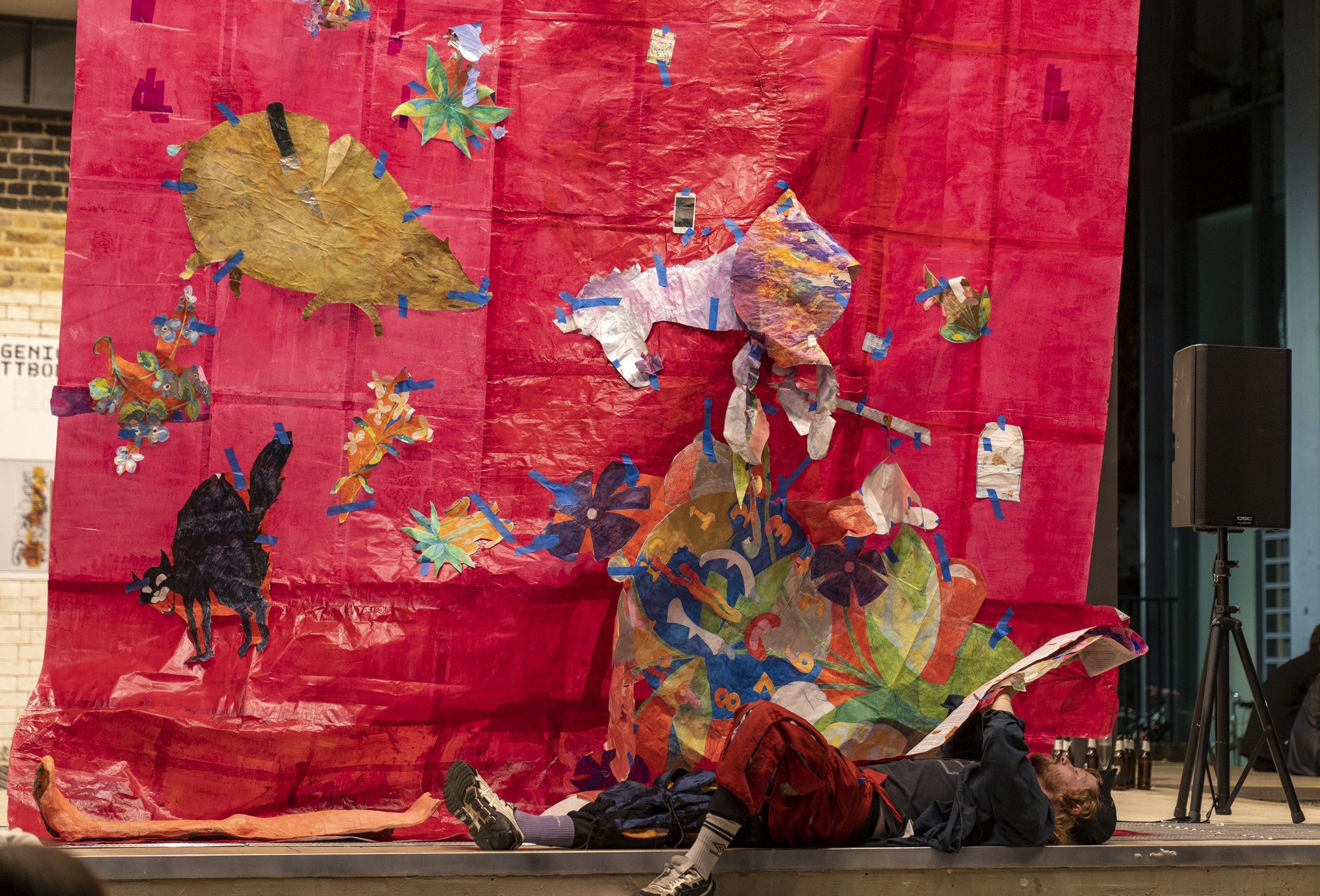 A white red haired man wearing landsknecht trousers and a baseball cap lays on his back reading a script. in the background, a large red backdrop made from paper, with drawings of a pig, a unicorn and a woried cartoon clock taped to its surface