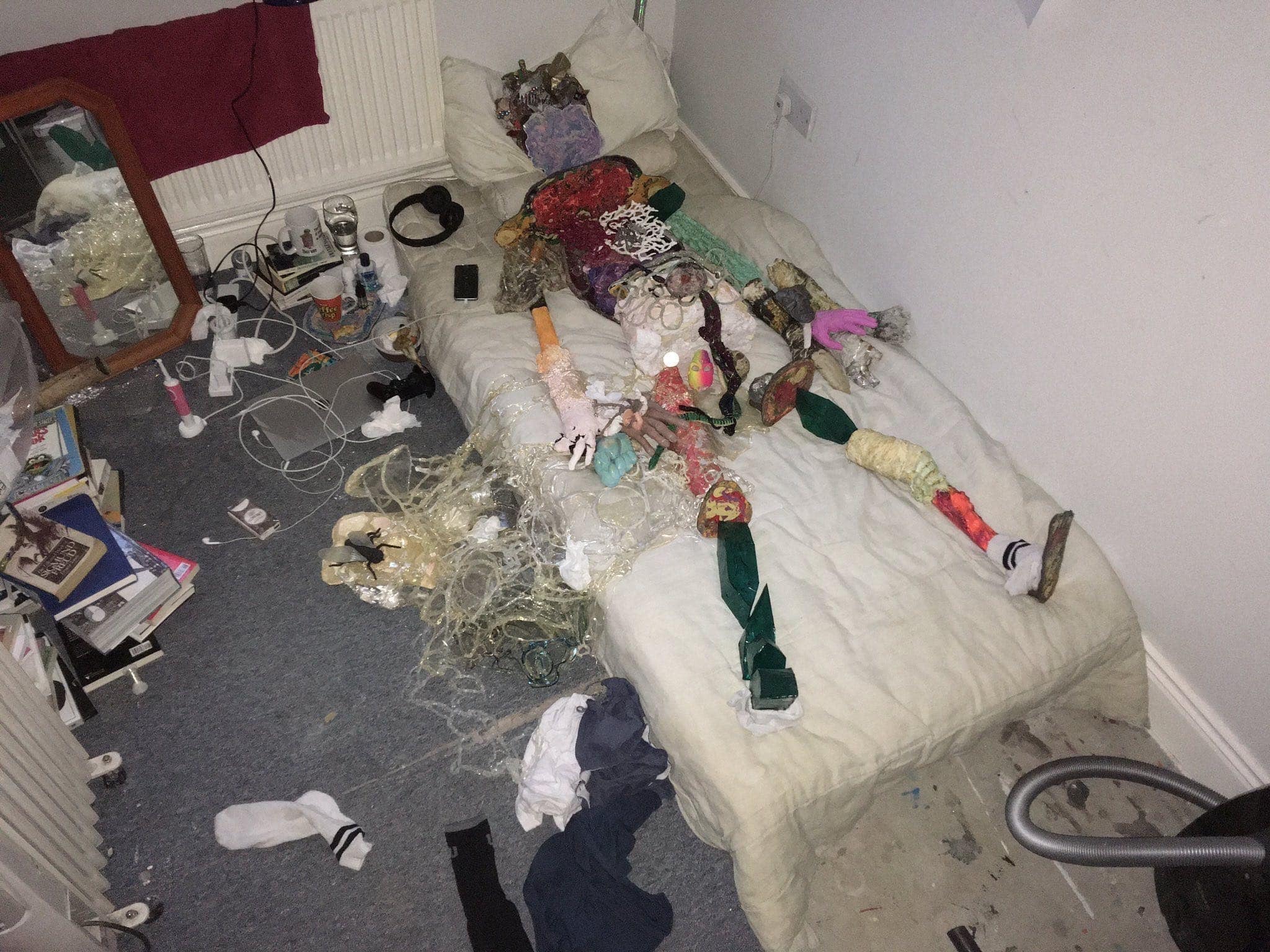 A figure is seen from above, laying on a matrass on the floor of a messy bedroom. The figure is made up of lots of small, shiny colourfull sculpture and the bed looks grimey, without sheets