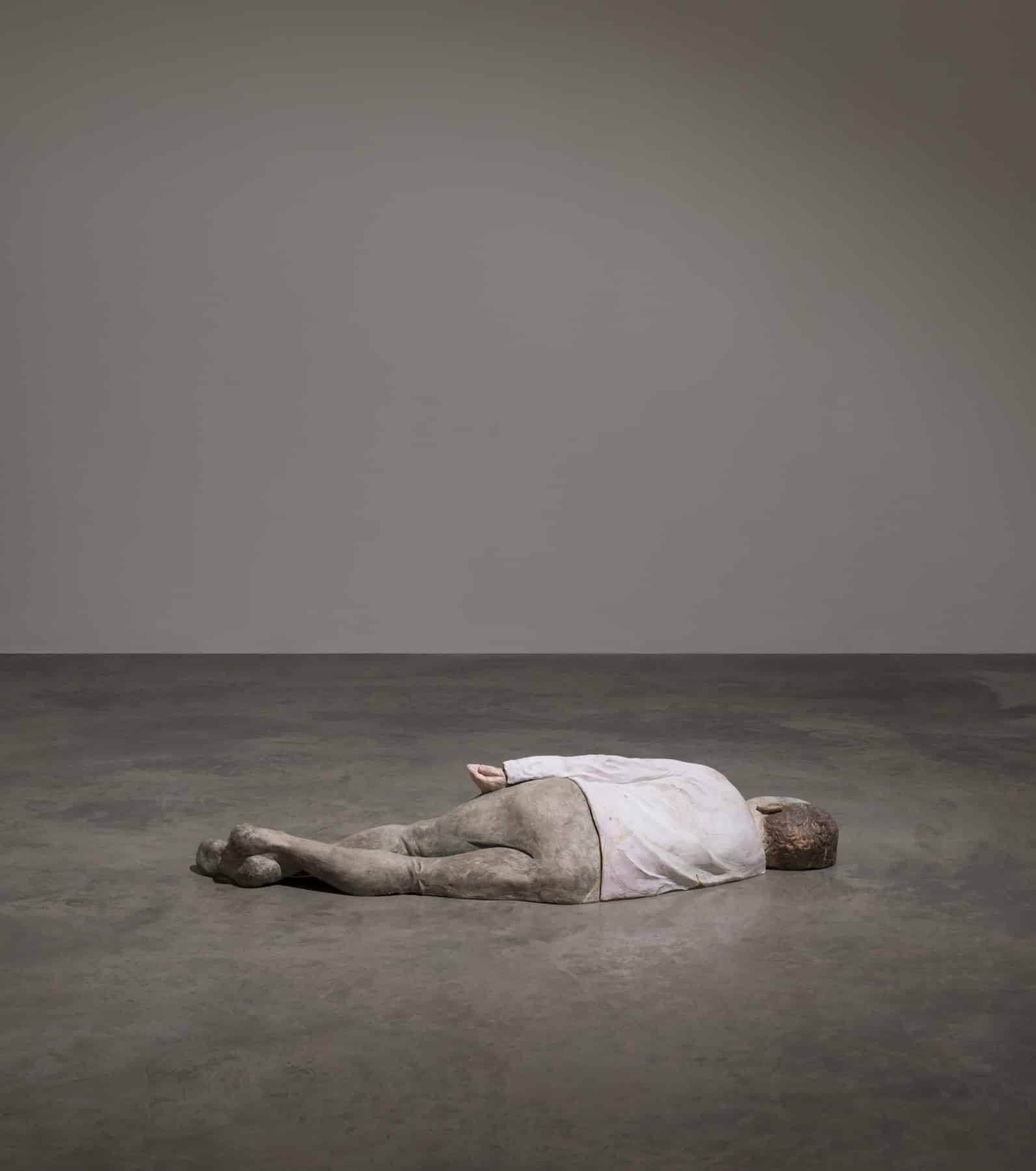 A figure is seen laying on its side on the ground. We see its back. They were a long sleeved white shirt and tight grey trousers.