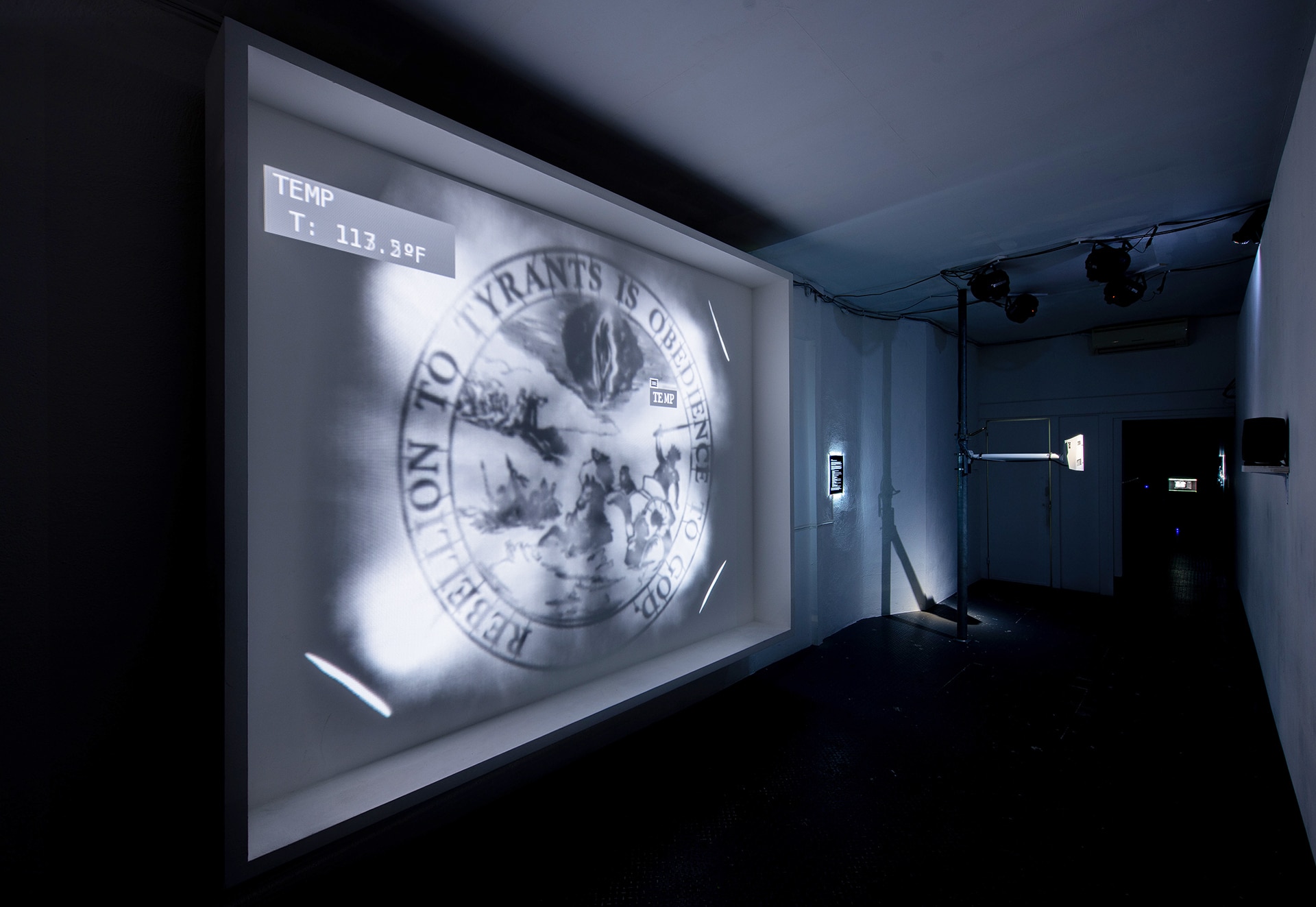 Installation in a darkened room showing a projected image from a thermal camera of a glowing white seal. First proposed and later rejected as the official seal of the United States, it depicts Moses crossing the red sea while pharoah and his armies drown in the water. Text on the seal reads: “Rebellion to Tyrants is obedience to God.”