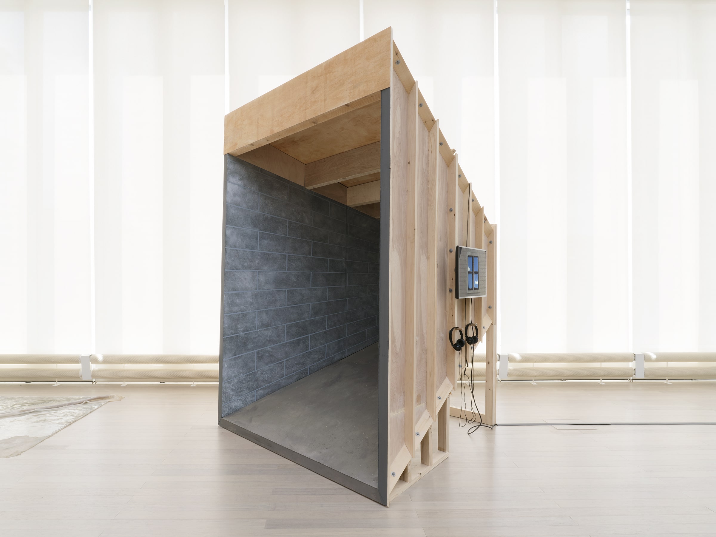 "Silver Bullet Test Site" overall shot. image description: A sculpture stands in front of a large row of windows (shades closed). The sculpture is a point perspective room constructed of wood and is painted to resemble a basement on the interior. a monitor hangs on the outside of the structure.