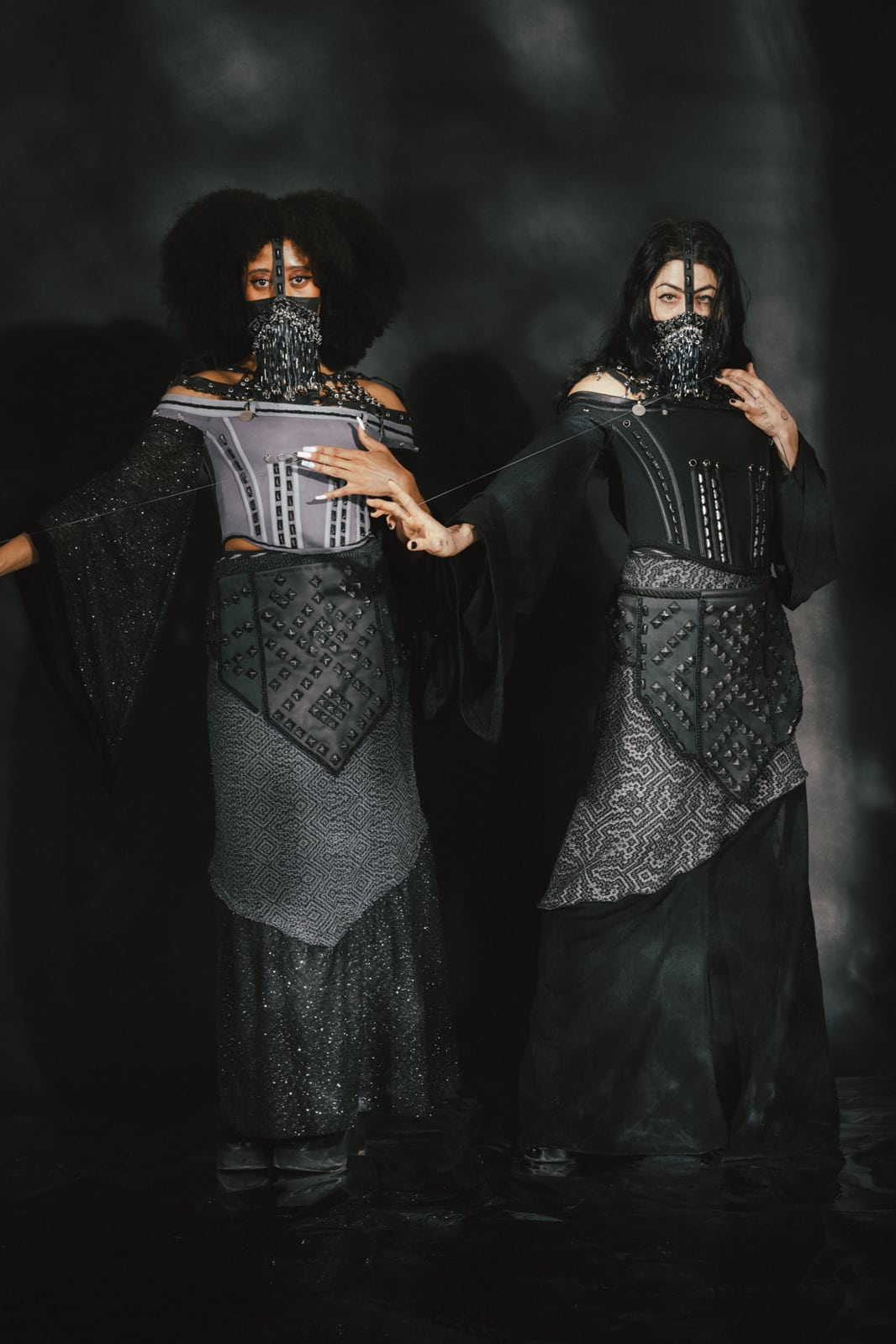 Two figures pose in front of an ethereally lit black and silver backdrop. Both figures wear variations of the same costume, including beaded face veils, a leather strap-spider web necklace with chain and beads, corset tops with beaded trim, and studded details, wide draped sleeves, handwoven skirts in repeating geometric patterns and leather paneled aprons, studded in patterns echoing geometric rug motifs. They are clad in all black and silver and both hold a thread taut between their hands.