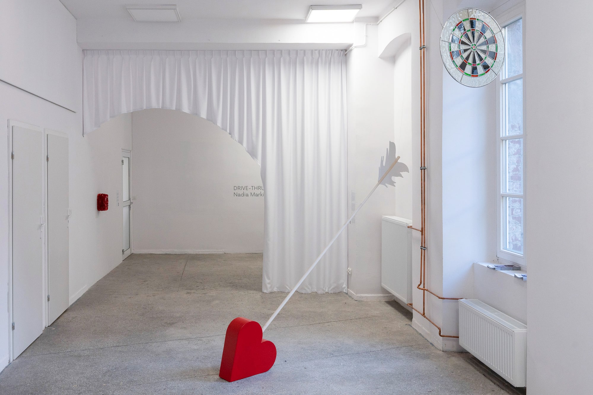 The image presents an interior with white walls and a gray terrazzo floor. On the wall on the right there are two white, closed doors. on the left hand side is a window. Towards the middle of the image, and room, are four objects. Starting from the left, on the wall next to the set of doors, in the distance, is a small box covered in a dark red, metallic festive garland. Right about above it, and crossing the room in half, facing the camera, is a white rippled curtain. A arch-like shape has been cut out in the curtain, making it possible to pass through. The arch is not in the middle of the square-shaped curtain, but a bit to the left of it, asymmetrically. In front of the curtain, diagonally cutting through the space and resting with its red, boxy arrowhead on the floor is a giant, wooden cupid's arrow. It has a light colored wood shaft and the feathers are a white wooden cut-out. Above the arrow, a bit to the right and overlapping the window, is a stained glass in the shape and colors of a dart board - beige, black, red and green.
