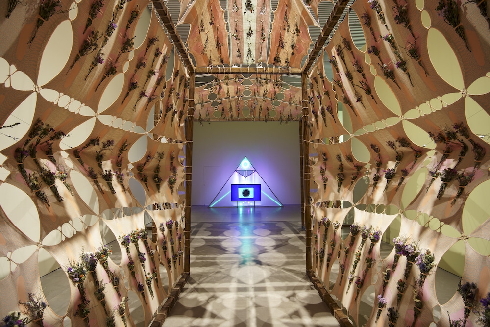 A maze of fabric and dried herbs leads to a neon-glowing pyramid strucutre.