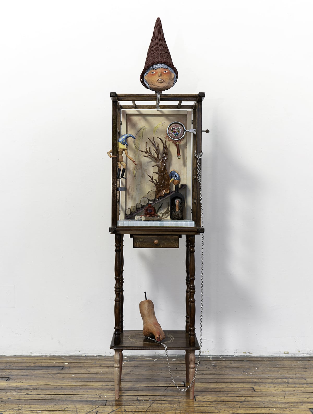 Assemblage sculpture on a wooden stand, featuring a doll-like head in a red hat, miniature figures, a barren tree, and chained objects.