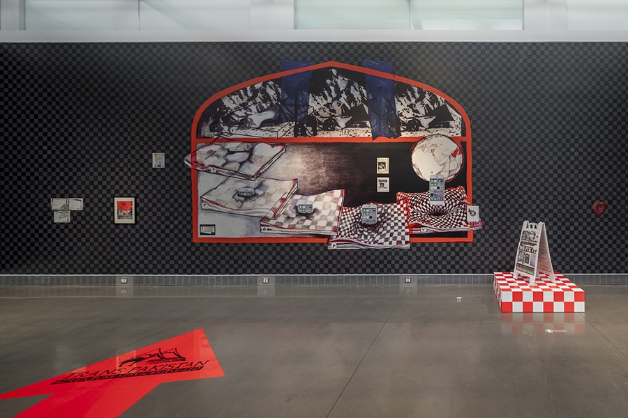A large red-outlined vinyl collage spans a checkered wall, featuring drawn maps, black mountains, and a globe. Nearby, a checkered pedestal holds a signboard.