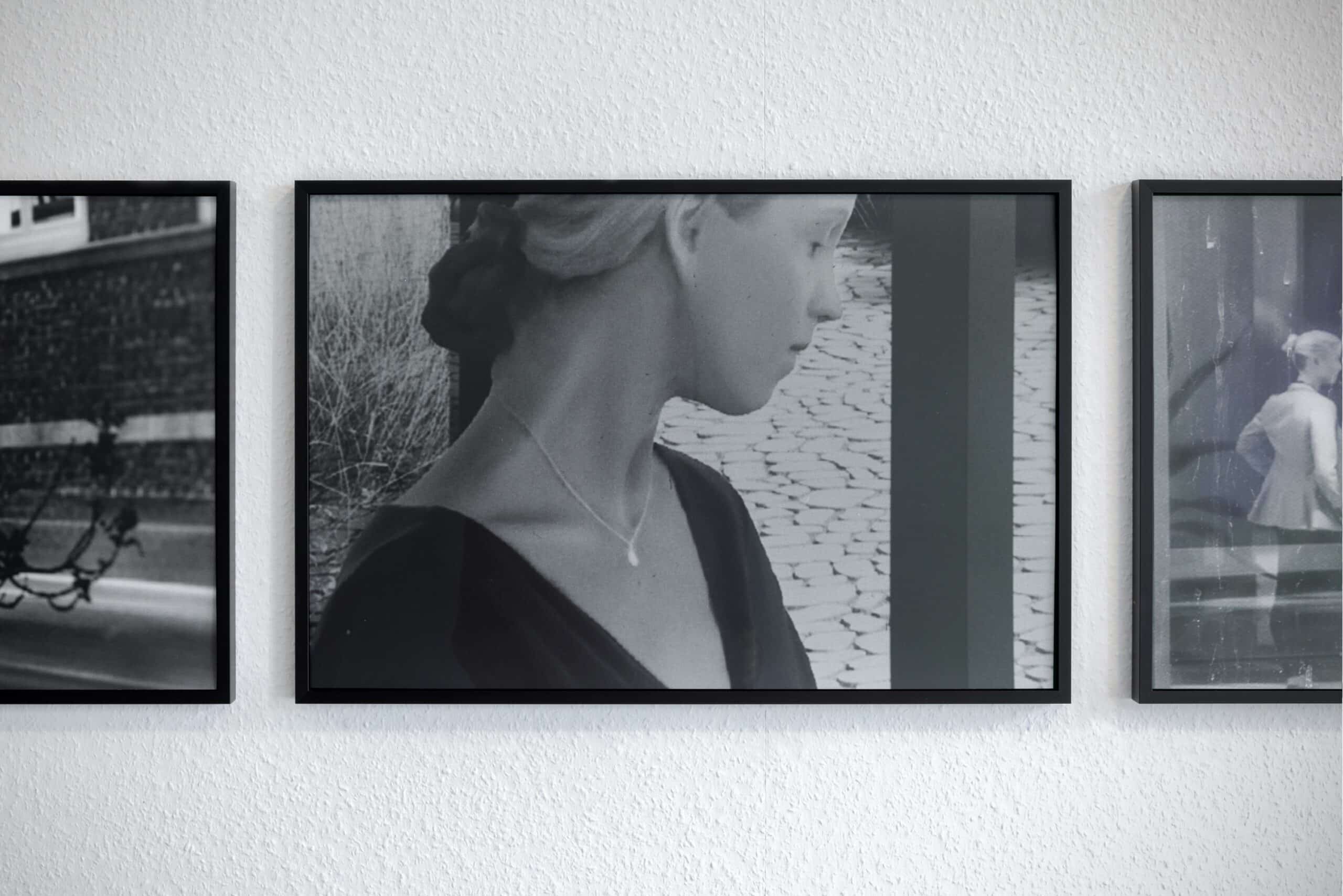 A framed black and white silver gelatine print of a woman's profile. She is looking away from the camera, her features feel uncanny, as if computer generated. The framed image is flanked by framed photographs in a similar style with just the edge of each image visible.