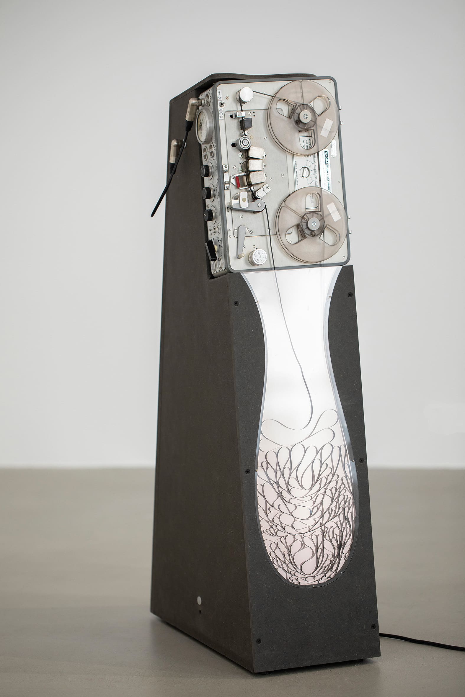 Meander: In a room with grey floor and white walls, there is a sound sculpture in which a Reel-To-Reel Tape Machine is integrated into a trapezoidal prism structure. Gravity causes the tape, emerging from the machine, to flow along a slanted incline into a bottle-shaped, flat, and luminous container made of Plexiglas. Within this form, the tape winds and gathers in meandering loops. In this way, a kind of drawing is created from the tape itself.