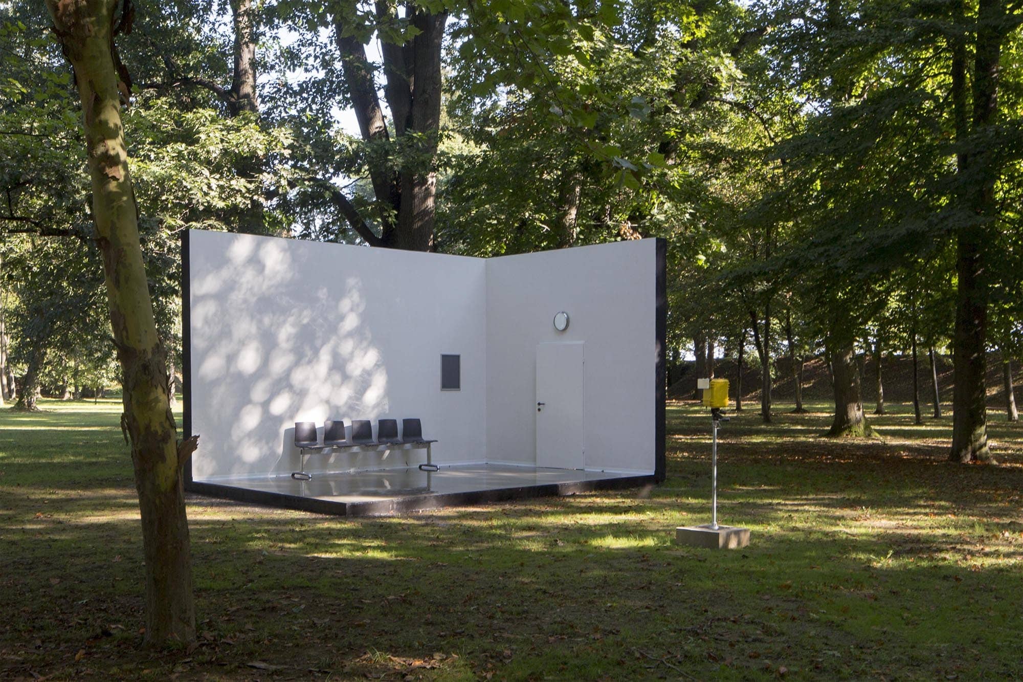 No Future: In a forest clearing, a room consisting of two walls and a concrete floor is visible. One wall has a closed door with a clock above it that has no face or hands, only a mirrored surface. On the other wall, an empty poster frame is mounted. In front of this wall stands a bench with five gray plastic aluminum seats. The arrangement resembles a waiting room but feels out of place in the middle of the forest. A camera in a weatherproof box is positioned in front of the room, aimed directly at it.
