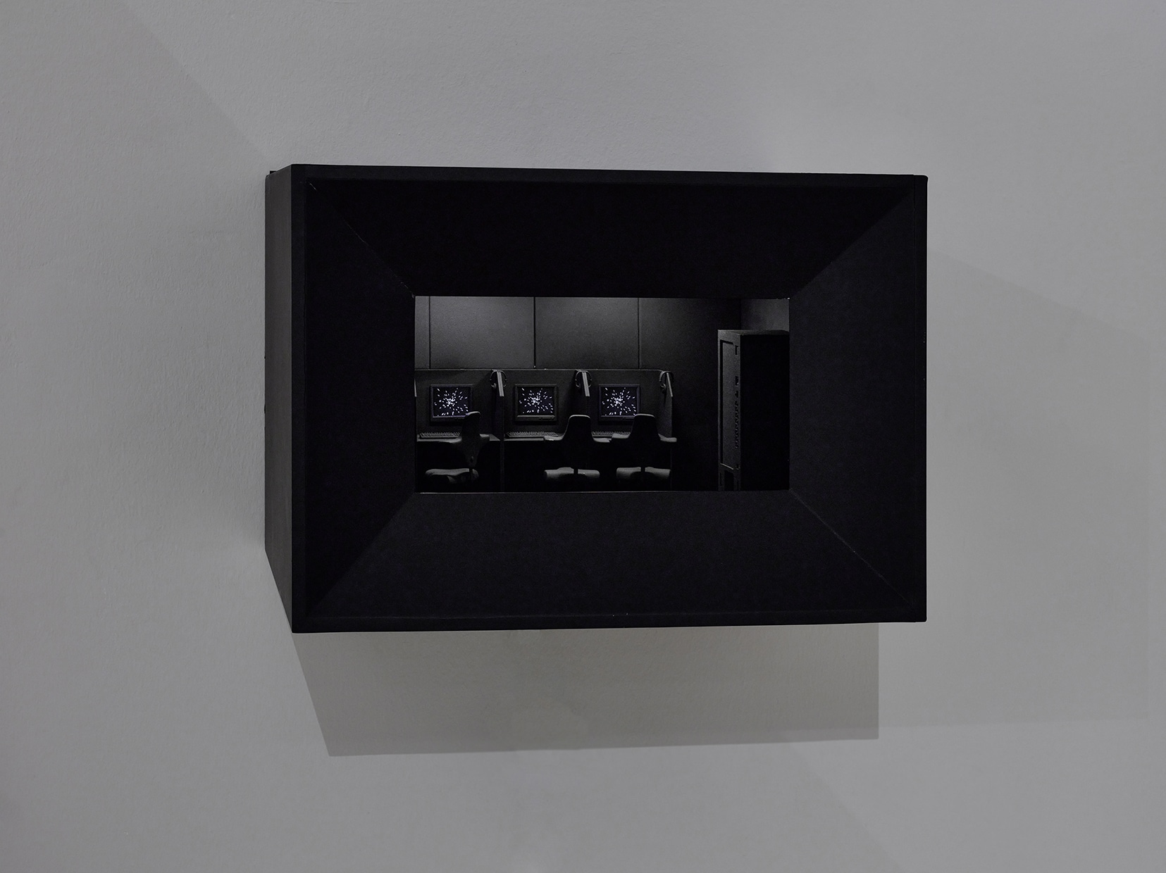 I looked around the internet: A black box protrudes from a wall. Through an opening in the box, a miniature room can be seen, containing three computer workstations. On the three desktop screens, screensaver animations of passing stars are running. The workstations are placed on a table, and are semi partitioned. Chairs are positioned in front of the workstations. To the right of the table, there is a vending machine.