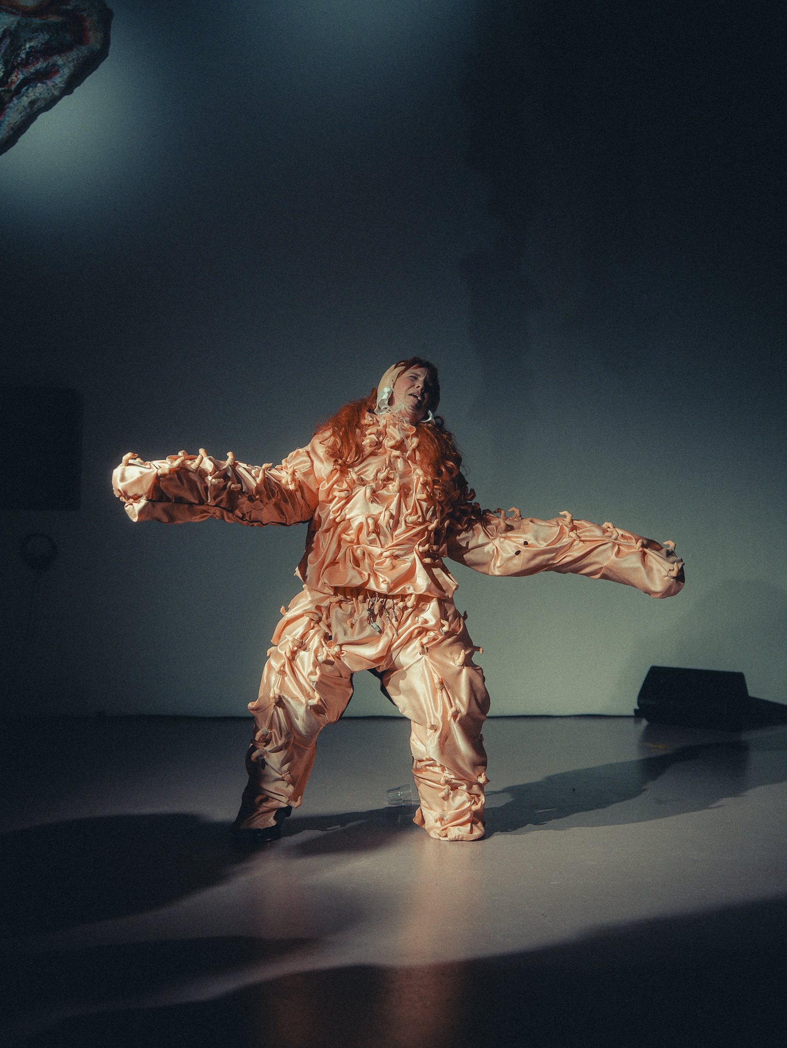 a fem robotic figure behind a white background covered ina oibnk satin suit and red hair. her face is framed by two plastic hands