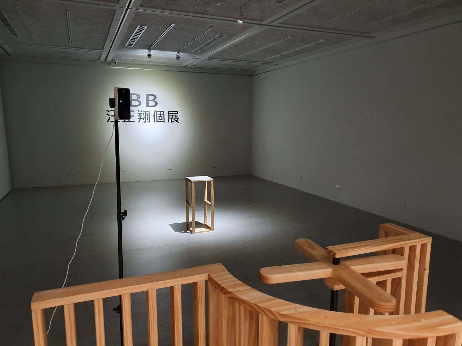 Installation view of a gallery space with a wooden turnstile barrier in the foreground. A small table stands under a spotlight at the center, while a smartphone mounted on a tall stand faces toward the barrier.