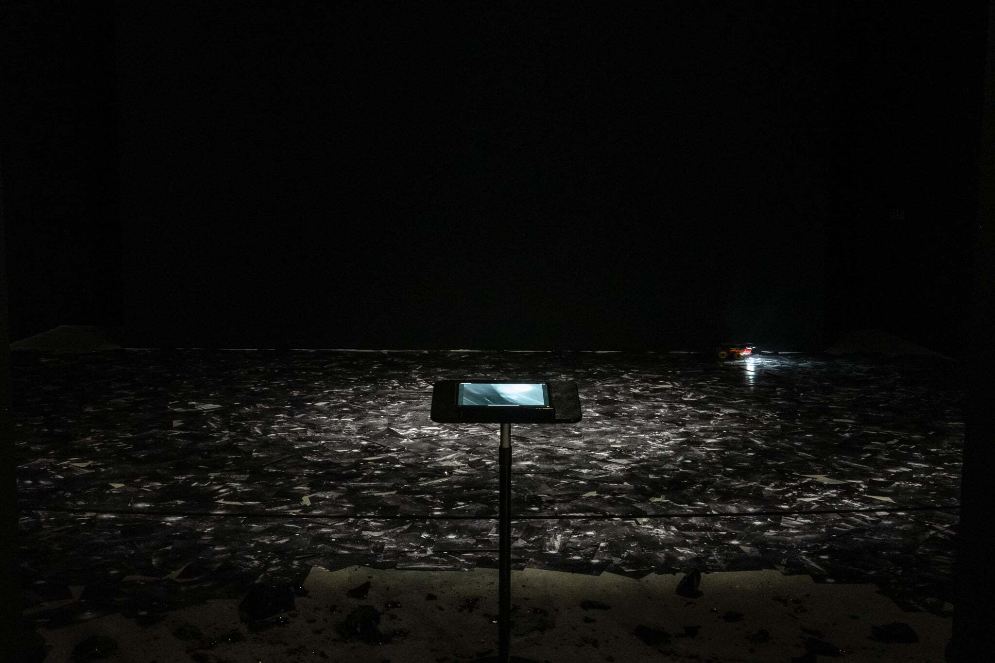 Installation view of a dark, frozen black lake-like surface made from photographic prints. A small remote-controlled car moves across the surface, projecting imagery onto an iPad mounted on a stand in the foreground.