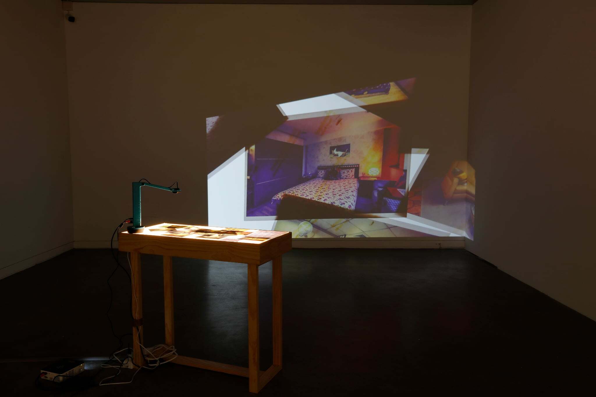 Installation view showing a wooden table covered with photographs under a desk lamp. Images from the table are projected onto a wall, forming a bedroom scene with a bed and a painting hanging above it.