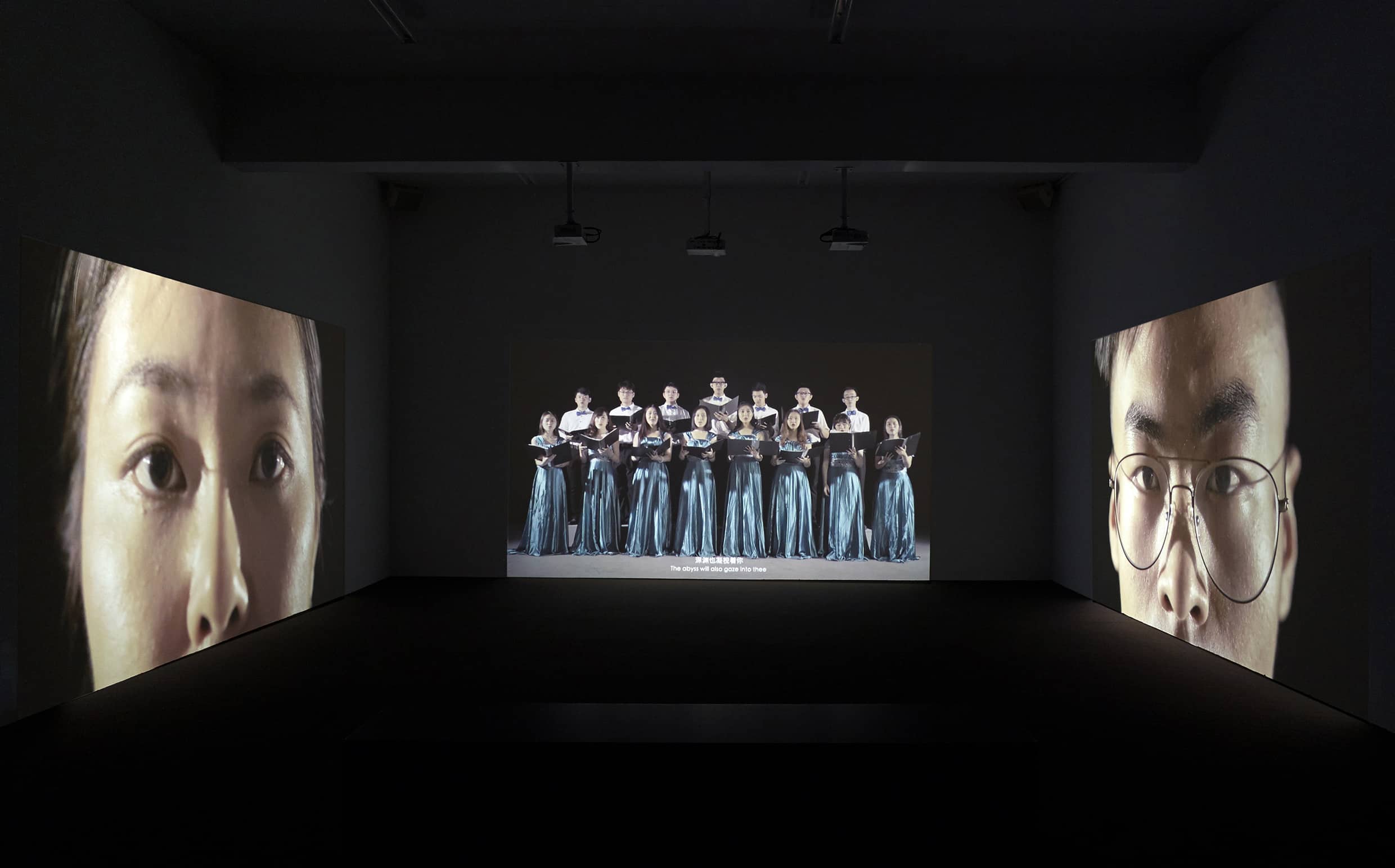 Three-channel video installation showing a dancer performing a burlesque striptease alongside a mechanical choreography, accompanied by a group of performers acting as a chorus. The work is presented across three screens surrounding the viewer, creating an immersive and staged viewing environment.